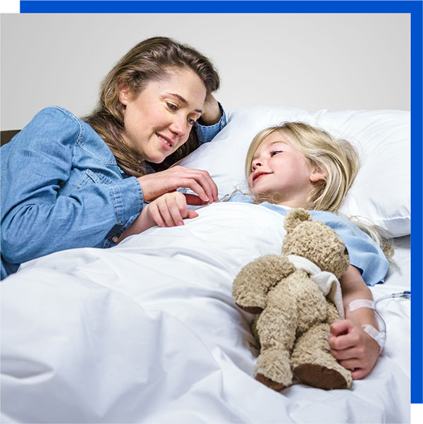 patient-and-family Photo of a woman laying next to a child in a patient bed. The child is in a patient gown with a stuffed bear in her arm.