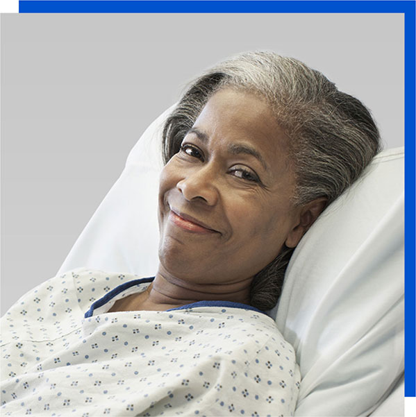 Photo of a smiling woman with gray and black hair wearing a patient gown and laying in a patient bed. 