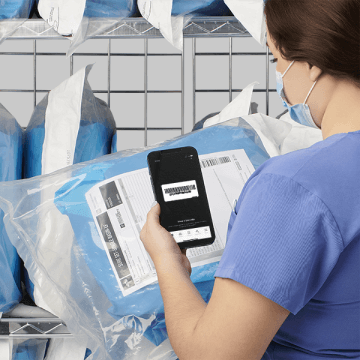 A tech in blue scrubs and a mask using an iphone to scan a barcode on a surgical pack.
