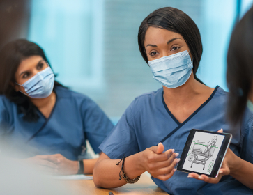 Masked nurse using a tablet to teach proper drain bag placement to other nurses