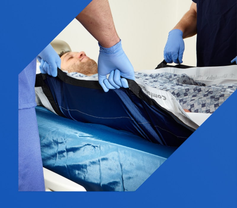 Two caregivers standing on either side of a hospital bed, using a repositioning sheet to move a patient