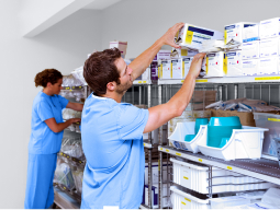 Male and female caregivers, wearing light green scrubs, pick supplies from shelves in storage room