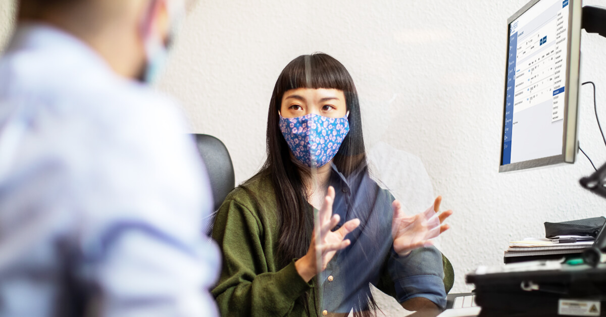 Woman with a mask at a computer talking to a man