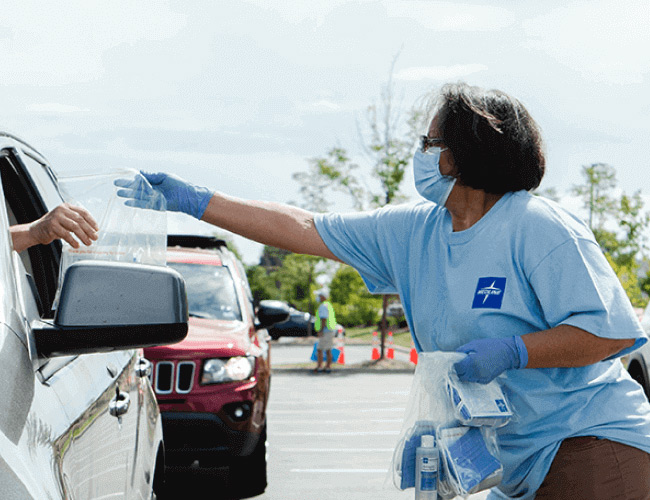 chicagoland-mb-bnr Medline employee in a mask hands plastic bag of supplies including hand sanitizer to woman in car in a parking lot.