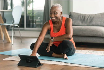smiling woman sitting on a yoga mat in her living room, looking at tablet propped up on the floor
