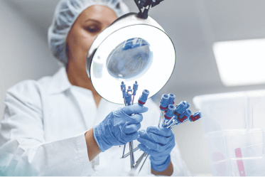 device-reprocessing woman wearing hair covering, lab coat and exam gloves inspecting single-use devices under a large magnifying glass