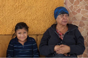 Older woman and young child standing against a stone wall outside.