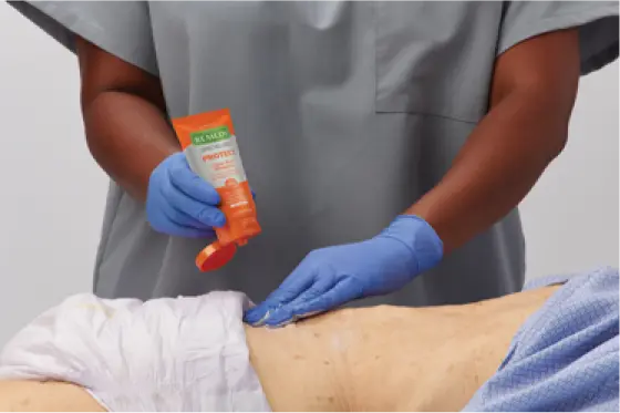 Gloved clinician hands apply Remedy protectant elderly person’s lower back by adult diaper..